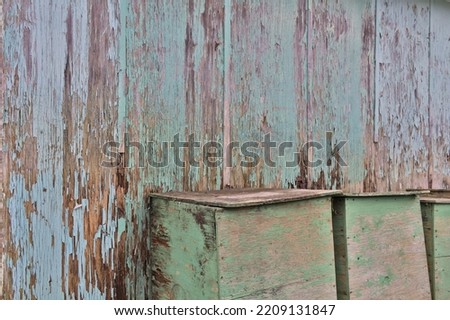 Paint chipped, weathered wood wall with wooden boxes in front of it. Aqua-green and aqua-blue paint chipping of and wood damp and rotting. Picture taken at an angle to the wall.