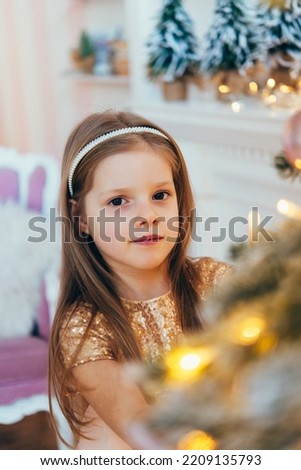 little girl, dressed in a beautiful fashionable white dress, poses near the Christmas tree. Beautifully decorated Christmas tree and fireplace, home interior.	