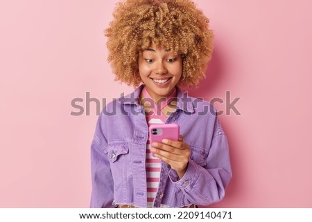 Indoor shot of optimistic woman uses modern smartphone for chatting online sending text messages smiles gladfully dressed in purple jacket isolated on pink background. People communication technology
