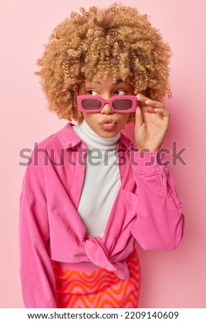 Photo of pretty thoughtful woman focused away keeps lips folded looks somewhere keeps hand on rim of sunglasses stylish shirt poses against pink background. Pretty female model with curly hair indoors