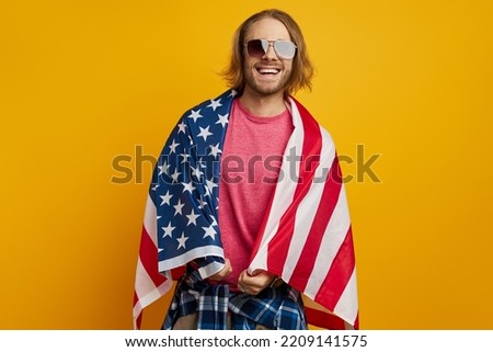Handsome young man covered with American flag looking at camera and smiling against yellow background