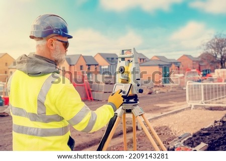 Site engineer in hi-viz working on house building construction site using modern surveying equipment against new houses nad blue cloudy sky background Royalty-Free Stock Photo #2209143281