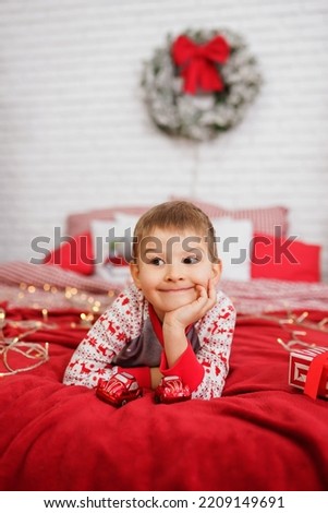 The European Boy is waiting for the New Year holidays lying on his stomach on the bed in red tones with a sad expression on his face. Emotions, the mood of the child on the eve of the holidays