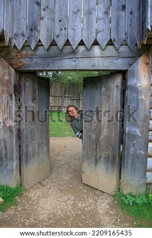 The photo was taken in the Carpathian Mountains near the entrance to a copy of a freely visited fortress. The picture shows a girl peeking out from behind an old wooden gate.