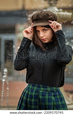 Portrait of a young beautiful dark-haired girl in a cap, a dark shirt and a plaid skirt in the city. Close-up.