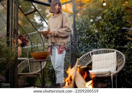 Young woman serves some food for dinner near picnic fire at cozy backyard with vintage glasshouse behind