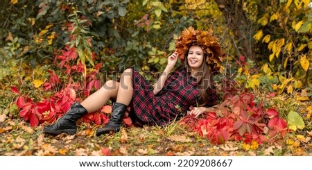 A happy smiling woman in a plaid dress lies in an autumn park against a background of colorful leaves. Artistic photo shoot with a girl in a wreath of maple leaves in the forest.