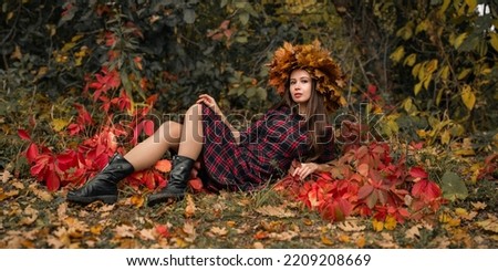A happy smiling woman in a plaid dress lies in an autumn park against a background of colorful leaves. Artistic photo shoot with a girl in a wreath of maple leaves in the forest.