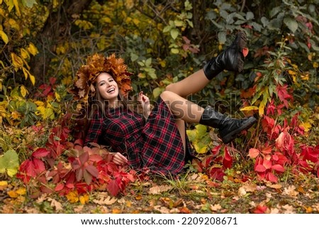 A happy laughing woman in a plaid dress lies in an autumn park against a background of colorful leaves and raises her legs. Artistic photo shoot with a girl in a wreath of maple leaves in the forest.
