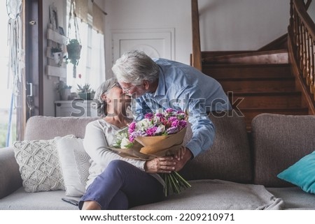 Old man giving flowers at his wife sitting on the sofa at home for the San Valentines’ day. Pensioners enjoying surprise together. In love people having fun. Royalty-Free Stock Photo #2209210971