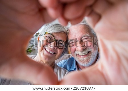 Couple of two old and happy seniors having fun at home on the sofa doing a heart shape with their hands and fingers looking at the camera. Royalty-Free Stock Photo #2209210977
