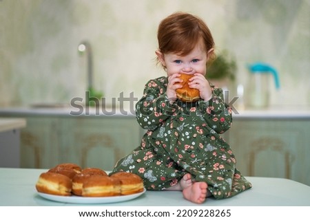 A little girl with red hair in a green dress sits on a table and eats sufganiyah donuts on Hanukkah day Royalty-Free Stock Photo #2209228265