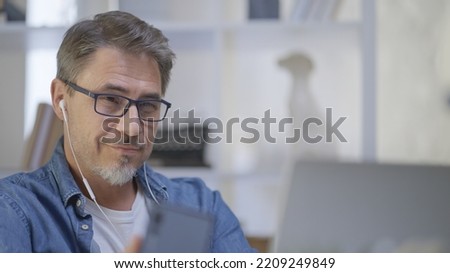 Older man sitting at desk in bright room working with laptop computer in home office. Mature age, middle age, mid adult casual man in 50s, serious, focusing on work. Royalty-Free Stock Photo #2209249849