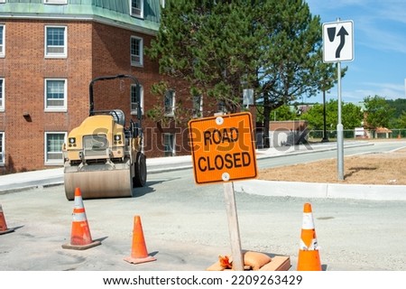 An orange colored road sign with road closed in black lettering. The sign is in the foreground with a road construction site in the background and a large brick building with tall green trees.