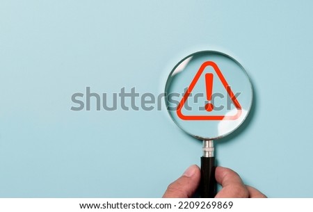 Hand holding magnifier glass with red triangle caution warning sign for focus notification error and maintenance concept. Royalty-Free Stock Photo #2209269869