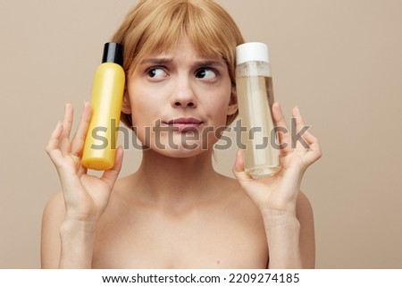 a funny, anxious woman stands on a beige background holding jars of care cosmetics in her hands and making an emotional face presses them to her head. Horizontal photo with empty space