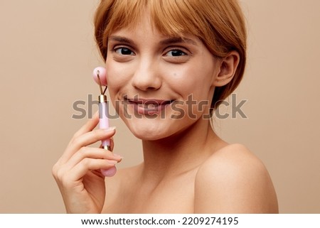 Horizontal studio shot.Close-up photo. A smiling woman in a towel with clean, beautiful, well-groomed skin, short red hair on a beige background does a facial massage with a new massage roller.