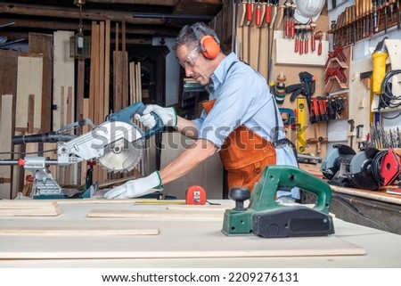 CARPENTER IN WORKSHOP WORKING WOOD WITH A MITER SAW. SMALL BUSINESSES AND SELF EMPLOYED. DIY AND PPE CONCEPT. PERSONAL PROTECTION EQUIPMENT FOR SAFETY AT WORK. Royalty-Free Stock Photo #2209276131