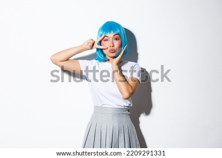 Image of flirty asian girl in blue anime wig showing peace gesture and winking at camera coquettish, standing over white background