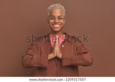 Young happy attractive African American woman joins palms in front of chest makes Namaste gesture symbolizing good intentions or appreciation dressed in casual clothes stands on brown background