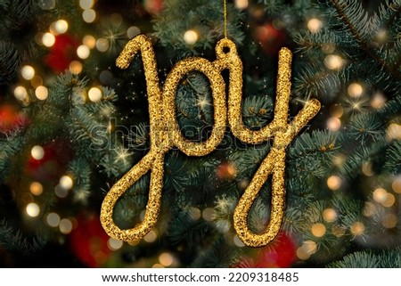 Christmas tree with gold ornament that says Joy, nestled in pine branches and lights. Holiday card.