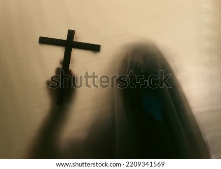 Horror ghost woman holding a cross behind the matte glass. Halloween festival concept.