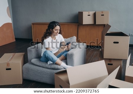 Hispanic girl uses phone app, searching moving company, shopping online for new home, sitting on armchair with carton boxes during packing things. Relocation, apartment renovation concept.