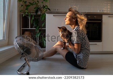 Young woman holds cat in front of electric fan and tries to cool down from stuffiness in apartment. Excited female sitting near fan helps pet to feel better from summer heat in flat kitchen Royalty-Free Stock Photo #2209382573