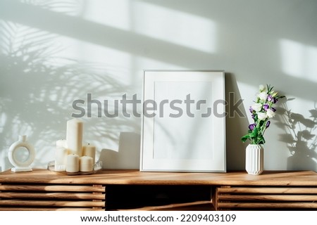 Modern minimalist Scandinavian style interior with white poster mockup, candles and flowers in vase on a wooden console under sunlight and home plants shadows on a white gray wall. Selective focus.