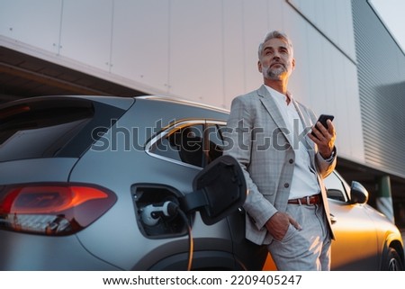 Businessman holding smartphone while charging car at electric vehicle charging station, closeup. Royalty-Free Stock Photo #2209405247