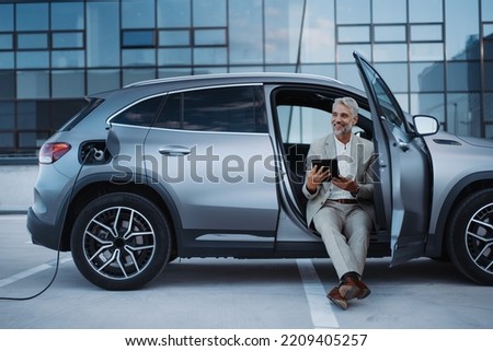 Businessman using tablet while charging car at electric vehicle charging station, close-up. Royalty-Free Stock Photo #2209405257