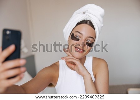 Beautiful young caucasian woman with towel on head and eyes patches takes selfie at home. Brunette hair female wears in white casual clothes takes care of skin. Concept gadgets, beauty.