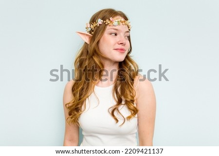 Young elf woman isolated on blue background looks aside smiling, cheerful and pleasant.