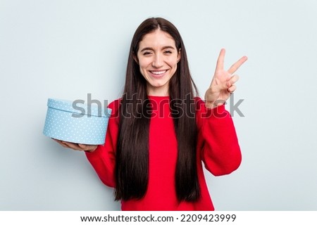 Young caucasian woman holding a gift box isolated on yellow background joyful and carefree showing a peace symbol with fingers.