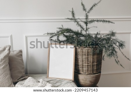 Moody Christmas still life. Blank vertical wooden picture frame mockup on sofa, linen cushions and blanket. Larch, pine tree branches in basket. White wall background. Elegant Scandinavian interior.