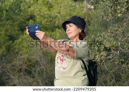 Elderly woman photographs the landscape during a hiking trip. A mature woman in panama and sportswear takes a photo with her smartphone. Concept of an active lifestyle of the elderly