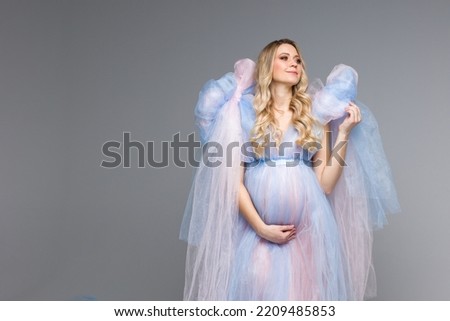 A pregnant belly. Pregnant girl with blonde hair on a gray background. Dresses for pregnant women. Maternity clothes. Vertical photo. Angel in blue dress. Copy space. Space for text.