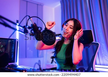 Asian young audio DJ woman sing a song on microphone to broadcasting. Attractive beautiful female blogger influencer wearing headphones and recording morning news podcast show for radio at home studio