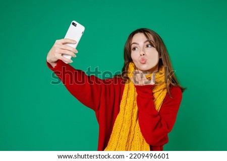 Pretty young brunette woman 20s wearing basic knitted red sweater yellow scarf doing selfie shot on mobile phone blowing sending air kiss isolated on bright green color background studio portrait