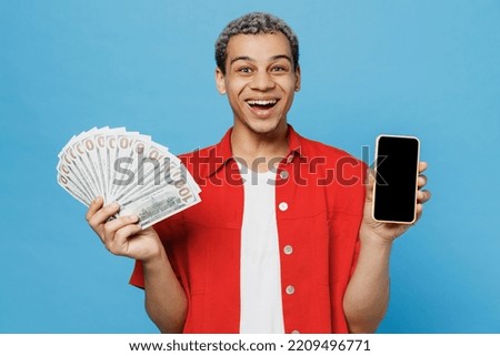 Young man of African American ethnicity wear red shirt hold fan of cash money in dollar banknotes use mobile cell phone blank screen workspace area isolated on plain pastel light blue cyan background