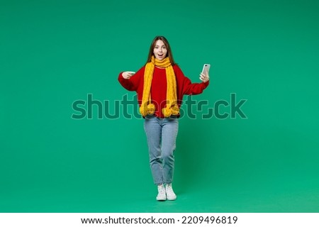 Full length of shocked young brunette woman 20s in knitted red sweater yellow scarf pointing index finger on mobile cell phone typing sms message isolated on green color background studio portrait