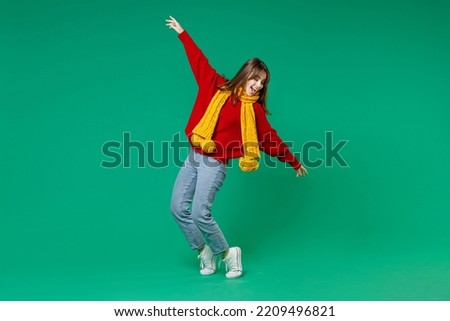 Full length side view of funny young brunette woman 20s in casual knitted red sweater yellow scarf dancing standing on toes spreading hands isolated on bright green color background studio portrait