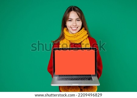 Smiling cheerful young brunette woman 20s wearing basic knitted red sweater yellow scarf hold laptop pc computer with blank empty screen isolated on bright green color wall background studio portrait