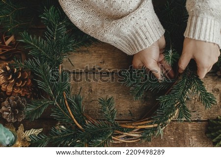 Making Christmas rustic wreath. Woman hands holding fir branches and making wreath on rustic wooden table with ribbon, golden bells, top view. Moody holiday image. Winter holiday workshop