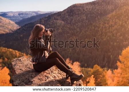 A young woman photographer in a fabulous autumn landscape in the mountains takes pictures on a professional camera, sitting on a rocky mountain top