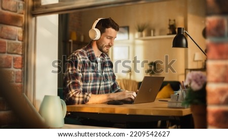 Modern Freelancer Wearing Headphones and Typing on Laptop, Creating Content for Social Media. Entrepreneur Working Remotely at Home Office, Managing e-Business. View From Outdoors into Window.