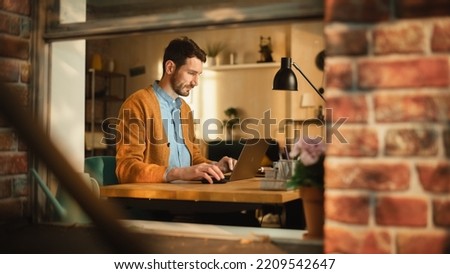 Handsome Man Typing on Laptop Computer in Loft Apartment, Sunny Day Outside. Creative Male Checking Social Media, Browsing Internet. Freelancer Working From Home. View From Outdoors into Window.