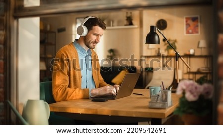 Thoughtful Man Using Laptop, Listens to Educational Audio Podcast in Headphones While Working Remotely at Home Office. Stylish Professional Doing Remote Work. View From Outdoors into Window.