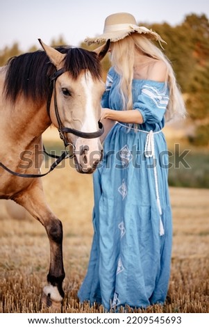 A young woman in a dress and hat walks her pet horse in a golden field in nature in the summer in the village