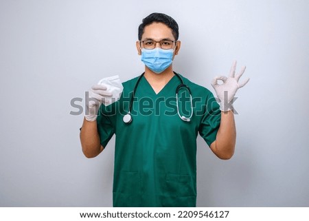 Cheerful smiling asian male doctor in scrubs holding medical mask suggest respirator while doing okay sign with finger, handing in to patient, white background